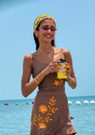 Woman in a brown dress with shell designs holding a yellow cup on a beach.