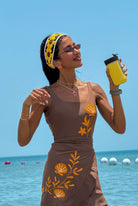 Woman in a brown dress with floral patterns by the ocean holding a yellow drink.